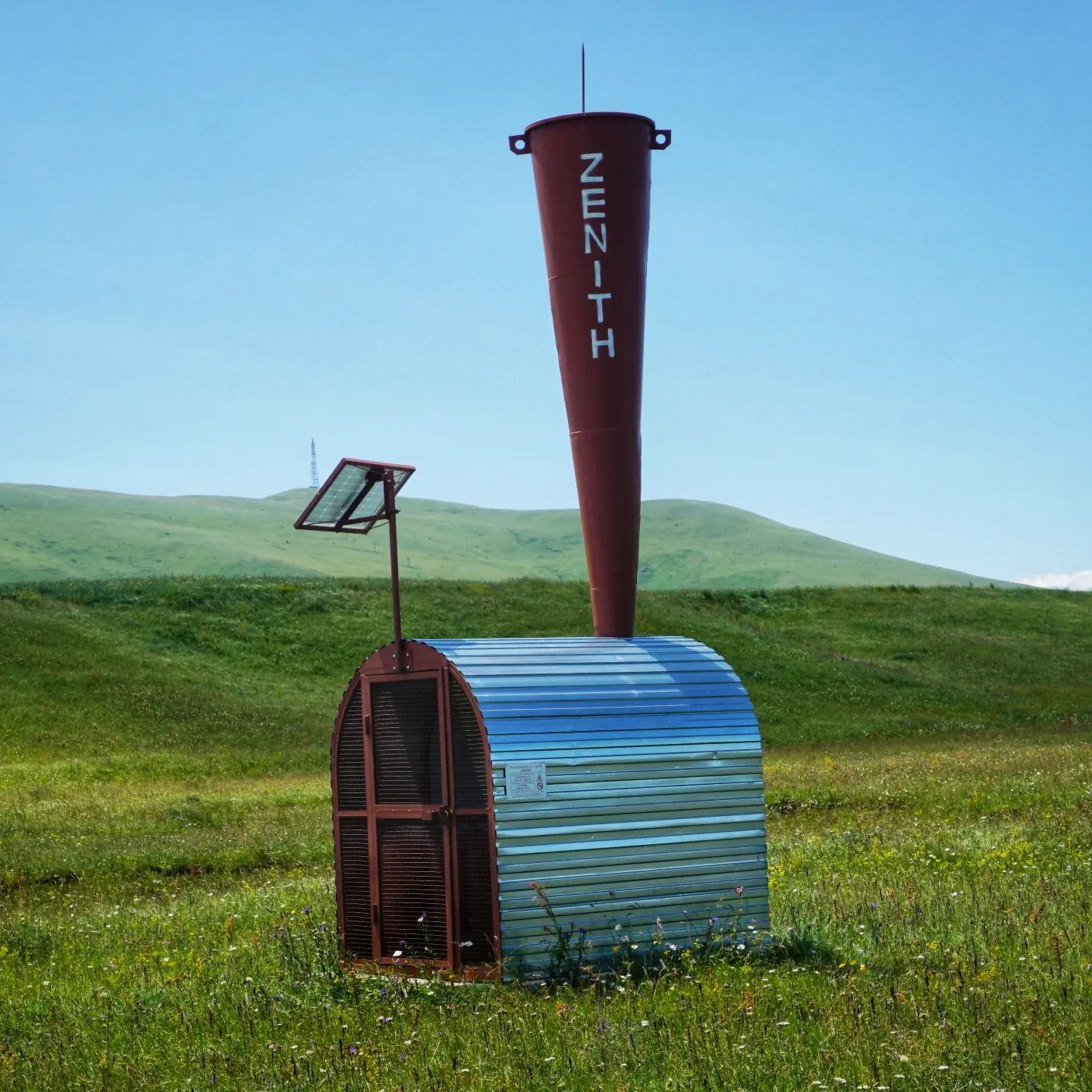 A photograph of a hail cannon, which is a small metal shed with a tall, upward-facing metal cone.